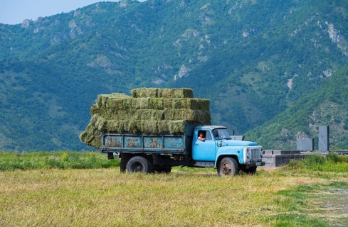 A blue truck with hay bales drives on a dirt road surrounded by green mountains, classic car,