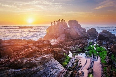 Scenic rock formation at Praia da Santa Cruz, Portugal, with ocean waves and vibrant sunset sky