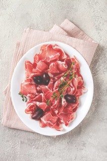 Freshly cut dried meat, served with olives and herbs, on a white plate, light background, no people