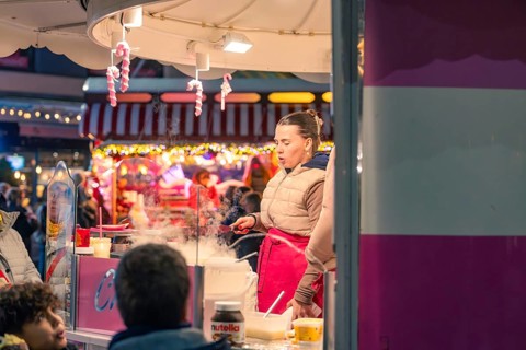 A woman cooks at a market stall surrounded by steaming food and colorful light, Christmas market