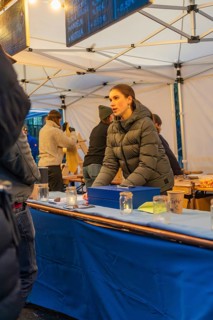 A woman in a winter coat sells goods at a market stall with a blue table, Aidlingen Christmas