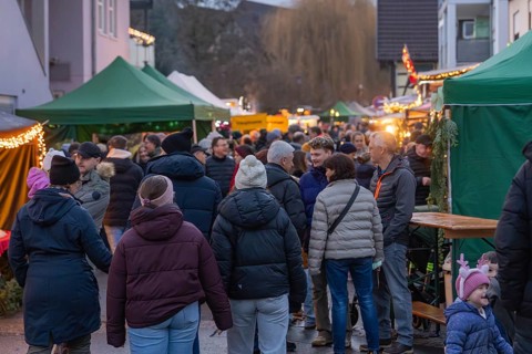 A crowd of people moves through a Christmas market with brightly decorated stalls, Aidlingen