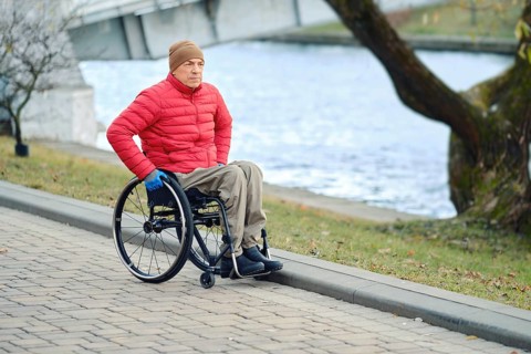 A man in a wheelchair moves along the river in the park taking a daily walk, wearing a red jacket