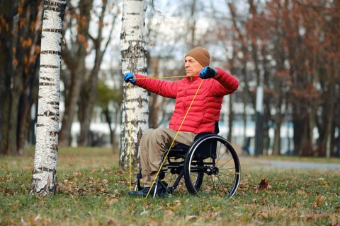 A man in a red jacket and beige hat performs strength training using resistance bands while seated