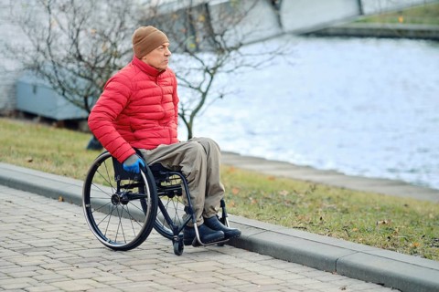 A man in a wheelchair stopped by a canal in a city park and looks at the water. He wears a red down