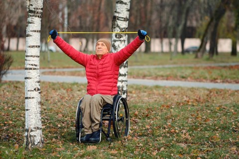 A senior old man sits in a wheelchair in a park, using a resistance band for exercise