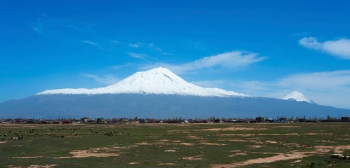 Snow-covered mountain range above a small village surrounded by green fields under a blue sky,