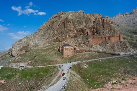 A fortress on a mountain with a road and cars in the foreground under a clear blue sky, Eski