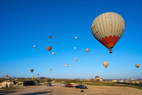 Several balloons float over a vast meadow landscape, with cars on the ground, hot air balloons,