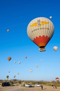 Hot air balloons rise from a meadow while people and cars are on the ground, Göreme National Park,
