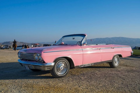 A pink vintage convertible car stands in a vast landscape with mountains in the background and blue