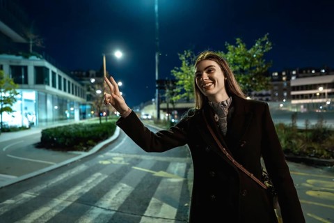 Young woman standing on an urban street at night, smiling and making a peace sign gesture while