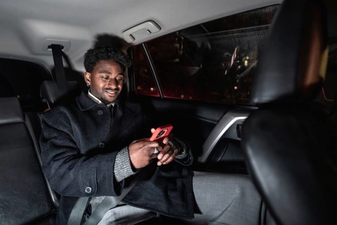 Young black man relaxed in a ride share backseat at night, smiling and browsing content on his