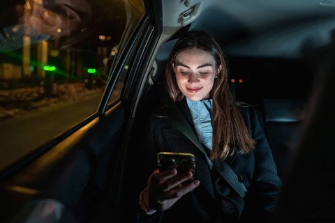 Young woman smiling while holding and looking at her smartphone in the backseat of a car at night,