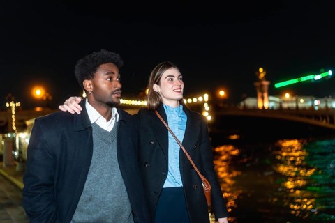 Young diverse couple strolling along a river promenade at night, city lights reflecting on the
