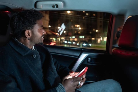 African american man in backseat of car at night smiling as he holds a glowing smartphone, looking