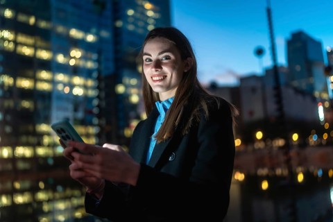Young woman texting on a smartphone, sharing a happy smile while standing on a city street at night