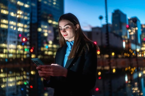 Young woman standing outdoors at night, using her smartphone with a glowing screen, surrounded by