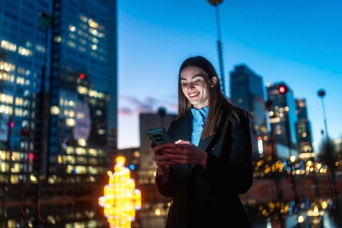 Young professional woman standing in a downtown night scene, smiling as her smartphone glows while