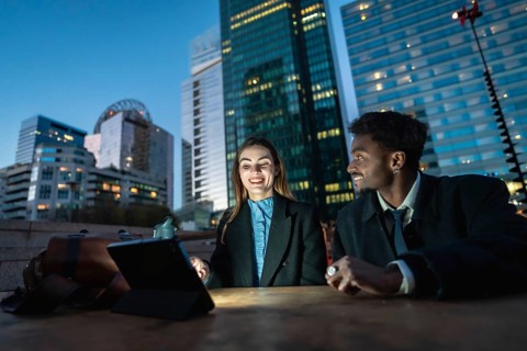 Two diverse business colleagues share a tablet at an outdoor table with a glowing downtown skyline