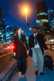 Diverse business couple walking with luggage along a downtown sidewalk at night, passing