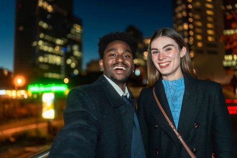 Diverse young man and woman smiling happily while taking a selfie together outdoors at night, with