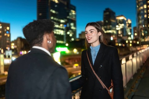 Two colleagues converse warmly on an urban pedestrian bridge at night, city lights and blurred