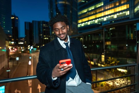 Young black businessman on an elevated walkway at night, smiling as he uses his smartphone with