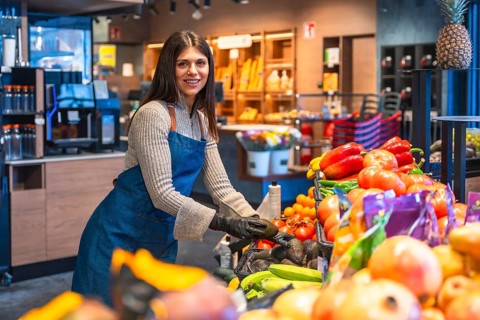 Woman in apron and gloves arranging fresh avocados on a supermarket produce display, smiling while