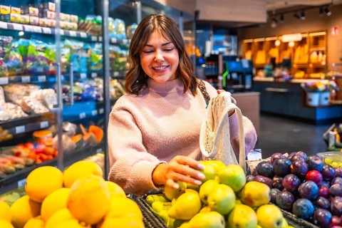 Young woman in a supermarket produce aisle selects ripe pears, carrying a reusable string bag for