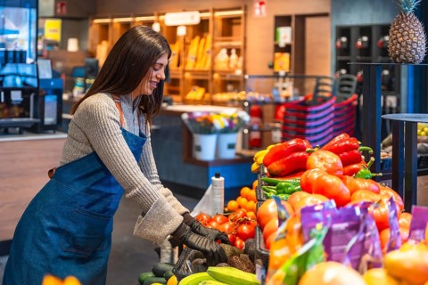 Happy young woman wearing an apron and gloves, organizing and stocking fresh fruits and vegetables