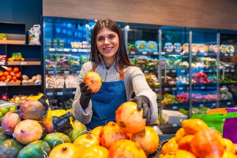 Smiling young woman greengrocer wearing an apron and gloves at a supermarket produce stand, proudly