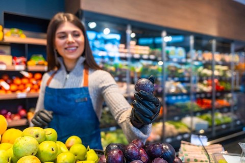 Smiling saleswoman in an apron and gloves working at a supermarket's produce section, holding a