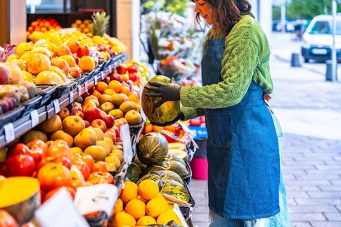 Female salesperson wearing a denim apron and gloves, arranging cantaloupe melons on a colorful