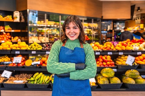 Smiling young greengrocer in blue apron and green sweater stands confidently with arms crossed amid