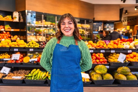 Woman wearing apron and green sweater standing with a friendly smile in a modern fruit and