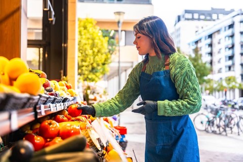 Young greengrocer woman wearing an apron and gloves arranging fresh organic fruits and vegetables