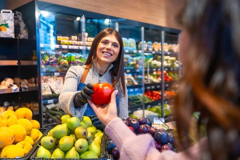 Friendly saleswoman in apron and gloves offering a fresh red bell pepper to a smiling customer in a