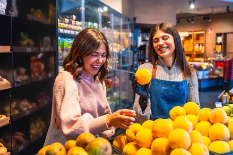 Female customer happily selects fresh oranges from a bright produce display while a cheerful apron