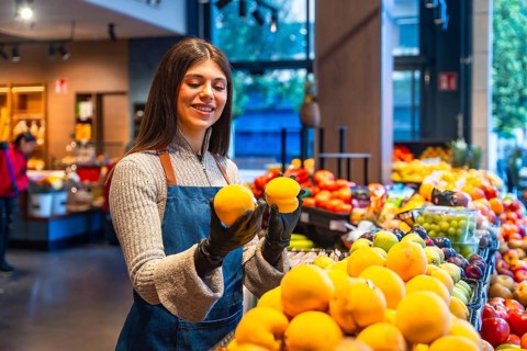 Produce worker wearing an apron and gloves arranging fresh oranges on a display, focusing on