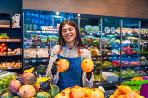 Female grocery store employee wearing an apron and gloves, smiling while holding two pomegranates,