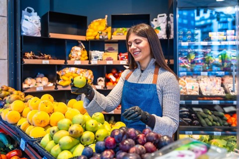 Smiling female greengrocer wearing an apron and gloves, carefully arranging fresh fruit on a