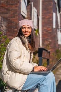 Woman in a cozy winter hat and jacket sitting outdoors, smiling while typing on a laptop, enjoying