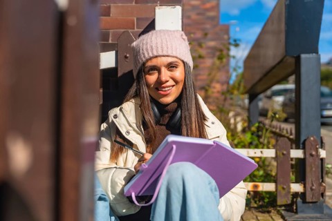 Young woman in beanie and headphones smiles while sketching in a notebook on a sunny urban street,