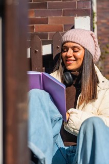 Woman smiling with closed eyes, wearing a beanie and headphones, sitting peacefully outdoors in the