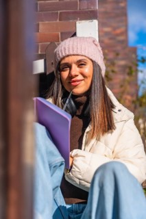 Woman student smiling and looking at the camera while writing in a journal outdoors, wearing a pink