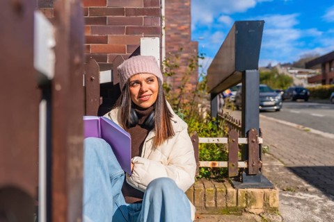 Young woman sitting outdoors on a sunny day, wearing a beanie and headphones, holding a book,