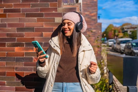 Woman smiling, wearing headphones and a beanie, holding a smartphone, standing against a brick wall