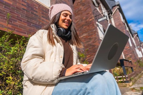 Young woman smiling and typing on a laptop, dressed in winter clothes, sitting outside a brick