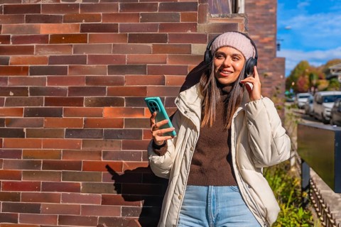 Young woman leaning against a brick wall, smiling with wireless headphones and smartphone, enjoying